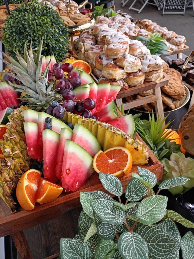 A vibrant fruit display on a wooden buffet cart, featuring sliced watermelons, whole pineapples, fresh orange slices, and dark red grapes, with pastries and other food items in the background.