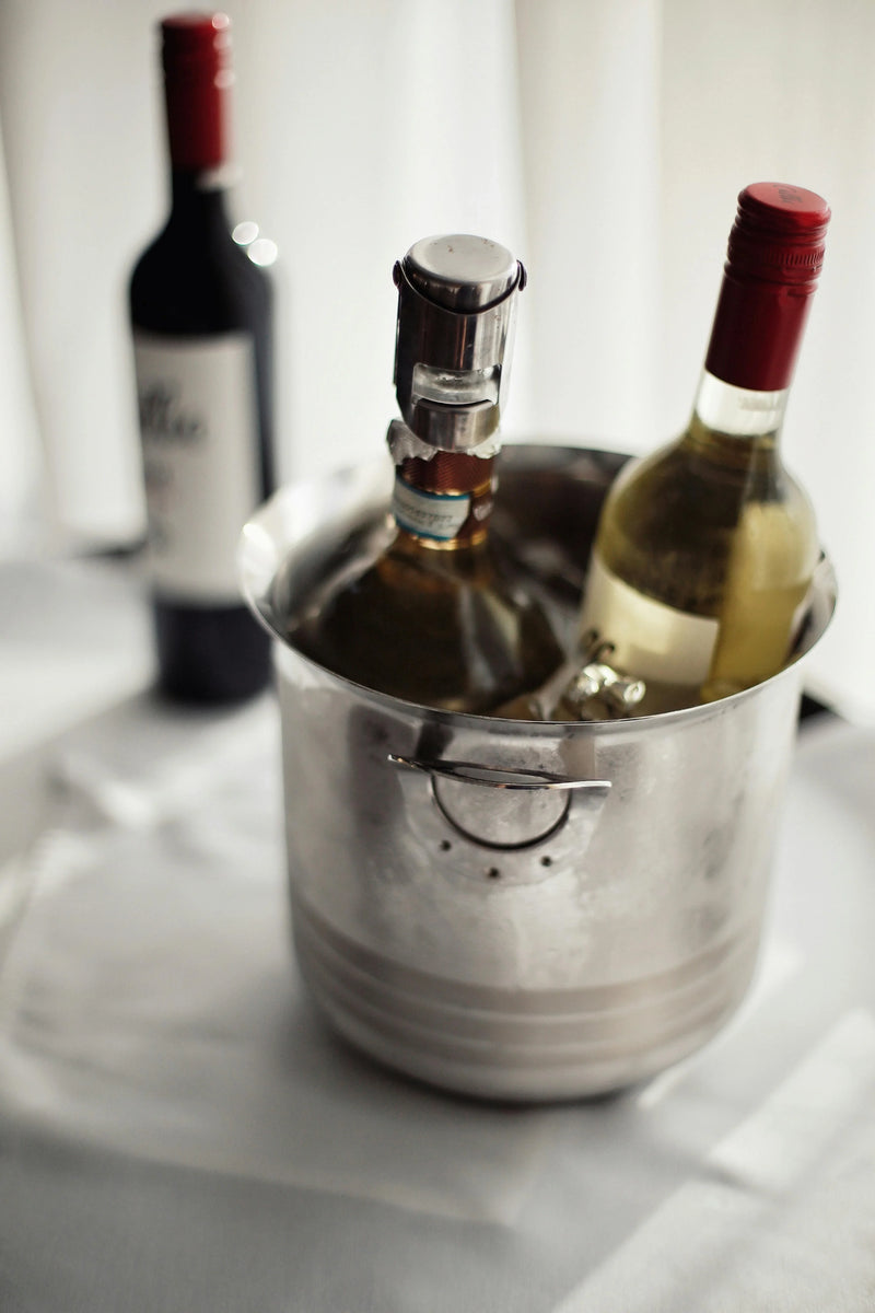 A stainless steel wine bucket filled with ice, holding two wine bottles, with a third bottle standing blurred in the background against a soft white curtain backdrop.