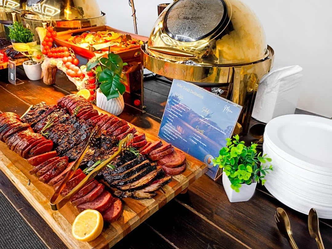 A buffet-style food display on a dark wooden table, featuring sliced grilled meats on a large wooden board, a gold dome chiller, white plates, small potted green plants, and a blue menu card.