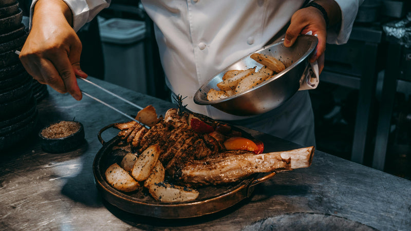 Person serving food from a metal bowl onto a dish with a large bone-in roast on a wooden table.