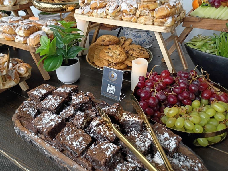A catering buffet table laden with assorted baked goods, including chocolate brownies dusted with powdered sugar, cookies, pastries, fresh red and green grapes, and a small potted plant on a dark wooden table.