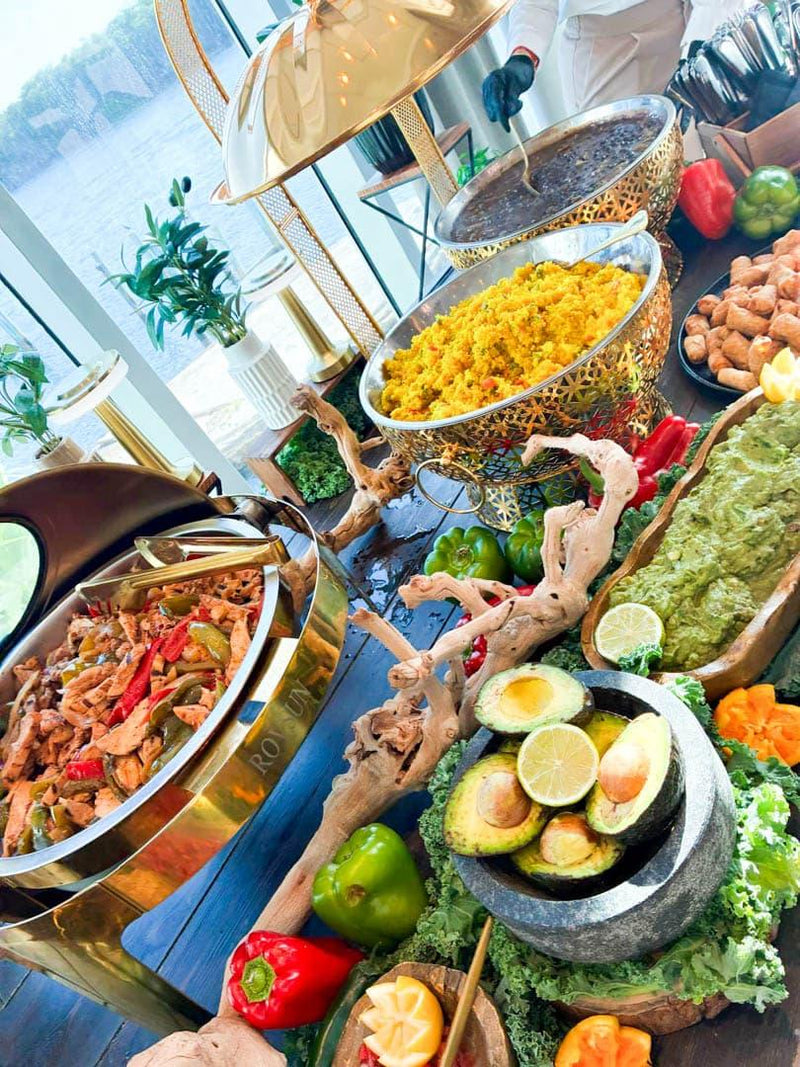 A buffet-style spread of prepared foods on a dark table, including a gold chafing dish of meat with peppers, a bowl of yellow rice, a green dip, sliced avocados, lime wedges, and vegetables, with decorative wood and brass serving pieces.