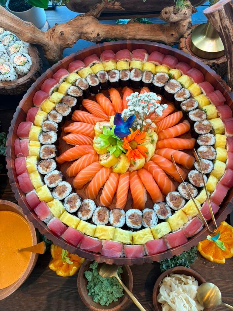 A large round platter of assorted sushi arranged in concentric rings, including salmon, tuna, yellow fish, maki rolls, and small garnish flowers, served on a dark wooden board with dipping sauces and side dishes.