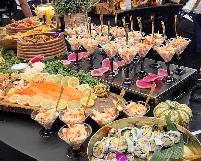 A buffet-style food spread with multiple glass cocktail glasses filled with shrimp cocktail, a large salmon fillet topped with lemon slices, assorted sushi rolls, and garnishes on a black serving table.