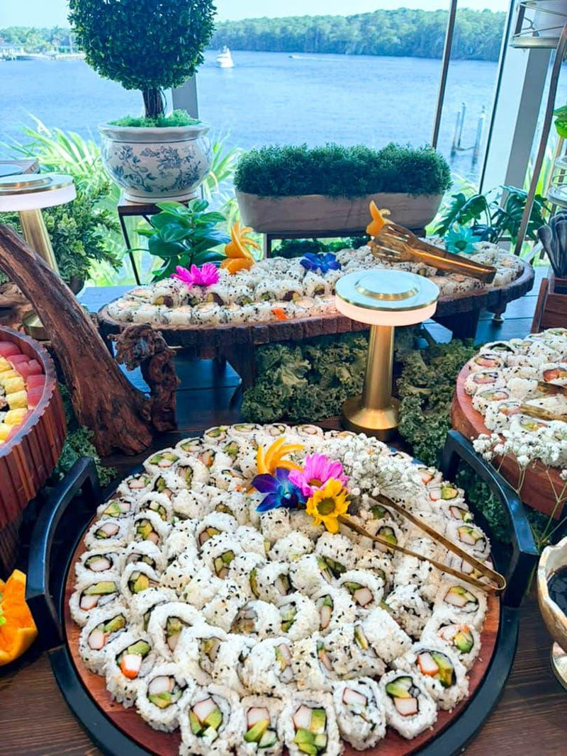 A large platter of assorted sushi rolls displayed on a buffet table, with a scenic lake and garden view in the background.