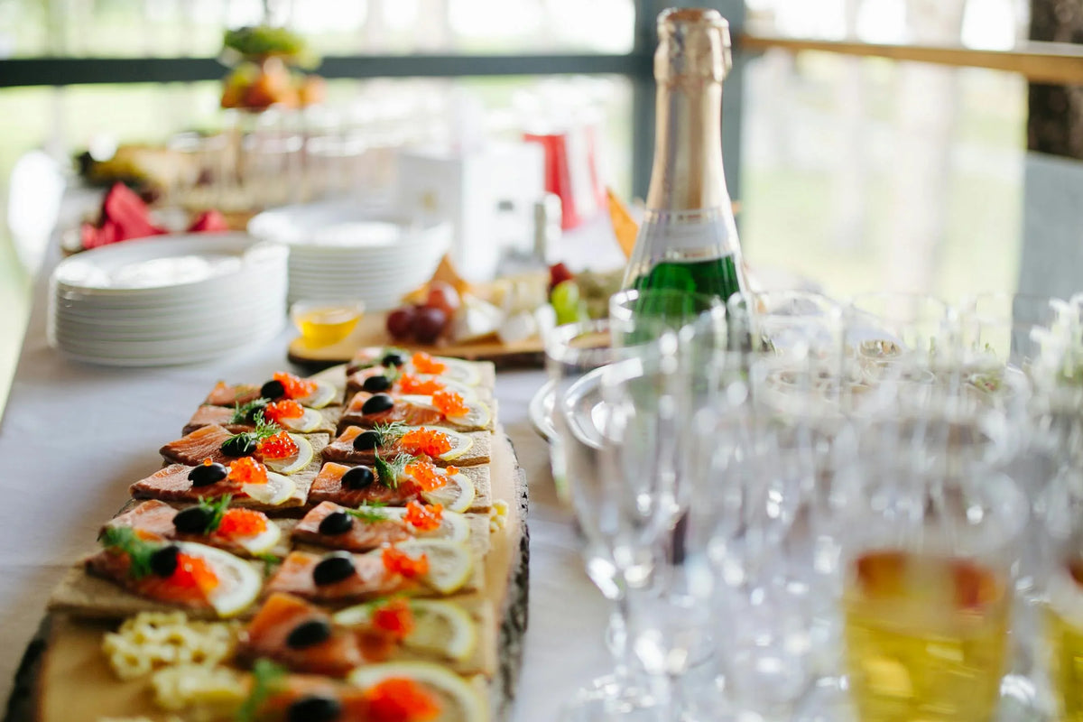 A catering table setup with a platter of appetizers topped with orange roe, black olives, and green garnish, alongside a bottle of sparkling wine, clear glasses, and stacked plates in a bright indoor setting.